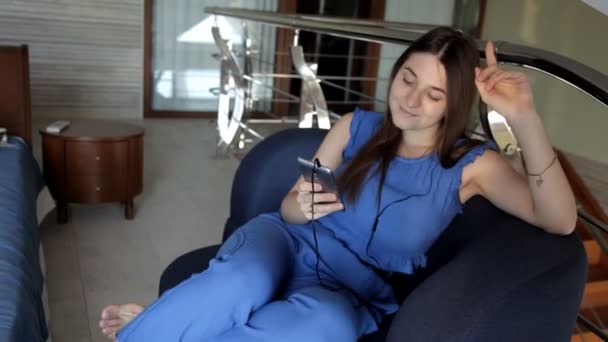 Молодая девушка сидит на диване, слушает музыку в наушниках и поет — стоковое видео