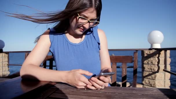 En flicka sitter på en restaurang på terrassen med utsikt över havet och läser ett SMS på telefonen — Stockvideo