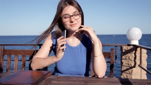 A menina se senta em um restaurante no terraço com vista para o mar, e o vento desenvolve seu cabelo e fala ao telefone. Sol e vento — Vídeo de Stock