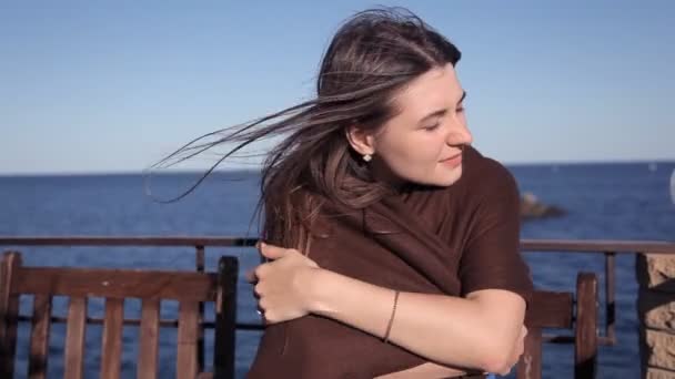 Uma menina senta-se em um café junto ao mar envolto em um cobertor soprando ao vento. Cabelo, vento, mar — Vídeo de Stock