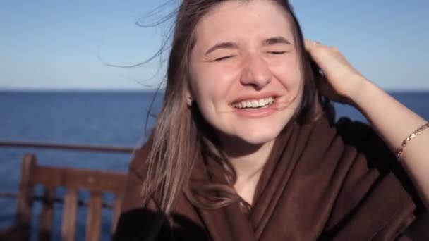 Una joven se sienta en un café junto al mar y se ríe. Un fuerte viento sopla en su cabello — Vídeos de Stock