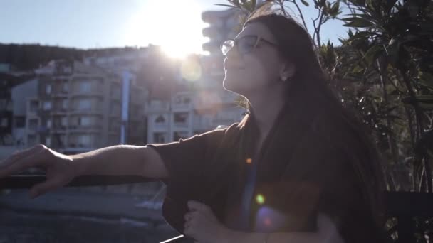 Η κοπέλα στο ηλιοβασίλεμα το βράδυ κάθεται στην βεράντα του καφέ με θέα τη θάλασσα και την πόλη. Ρομαντική βραδιά — Αρχείο Βίντεο