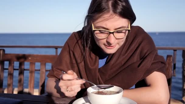 Дівчина сидить у вечірньому заході сонця сидить у кафе з видом на море, покритий ковдрою і їсть гарячий бульйон — стокове відео