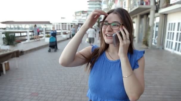 Una joven camina en una ciudad situada en la orilla del mar habla por teléfono y comparte sus impresiones — Vídeos de Stock