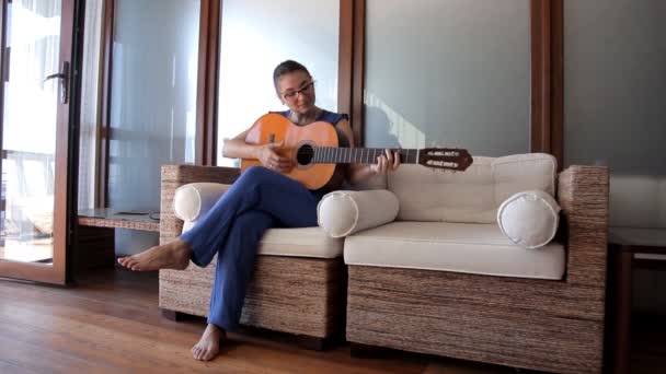 Una joven se sienta en el sofá y toca la guitarra — Vídeos de Stock