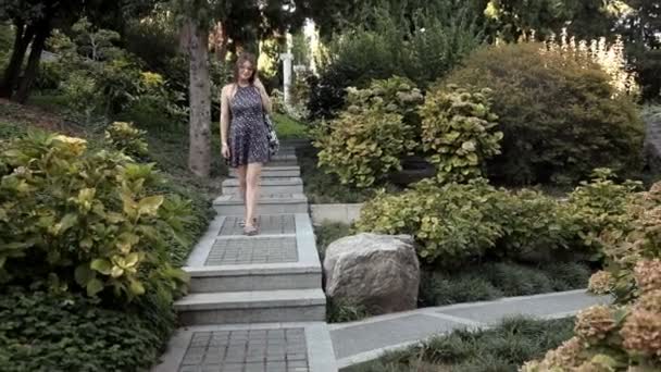 Seorang gadis muda datang menuruni tangga yang indah terletak di taman hijau dengan berbagai tanaman dan pohon aneh — Stok Video