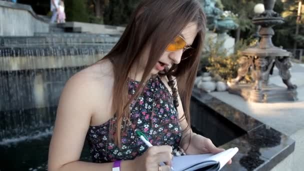 A young girl sits by the fountain and takes notes in a notebook — Stock Video