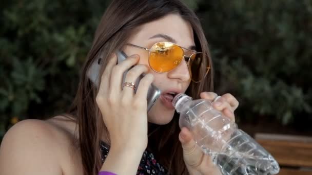 Una joven se sienta en un parque en un banco, bebe agua y habla por teléfono. Verano y naturaleza — Vídeo de stock