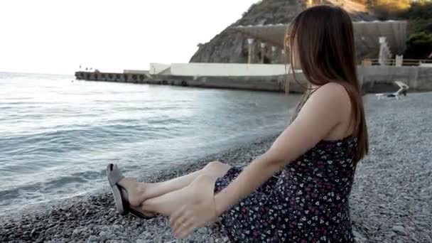 Una joven al atardecer, sentada junto al mar, escuchando música y cantando arroja piedras al mar — Vídeo de stock