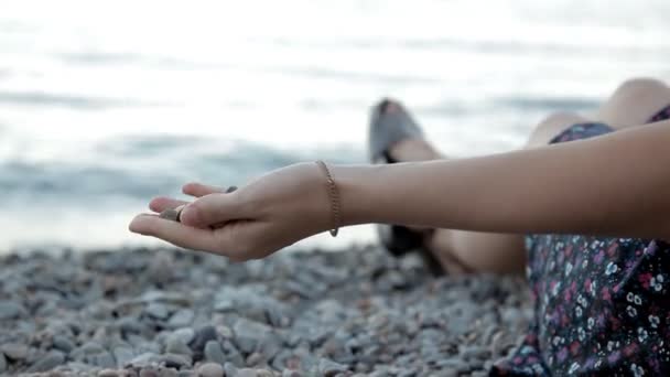 Une jeune fille au coucher du soleil est assise au bord de la mer et remet des pierres — Video