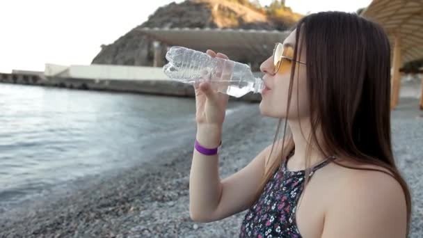 En ung flicka vid solnedgången sitter på stranden och dricker vatten. Sommaren berg — Stockvideo