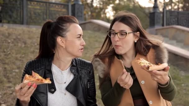 Две девушки сидят в парке и едят вкусную большую пиццу с курицей и грибами — стоковое видео