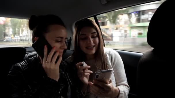 Dvě mladé holky v taxíku, jeden mluví do telefonu, a přítel jí ukazuje hudební video na její mobilní telefon — Stock video