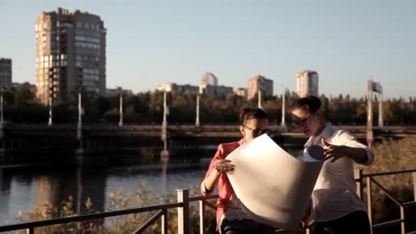 Два архитектора стоят на строительной площадке, на реке и рассматривают план строительства бизнес-центра — стоковое видео