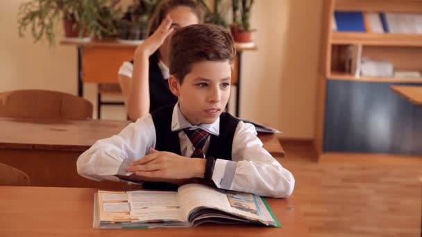 Pojke i uniform i klassrummet lyfter sin hand osäkert som skulle svara läraren — Stockvideo