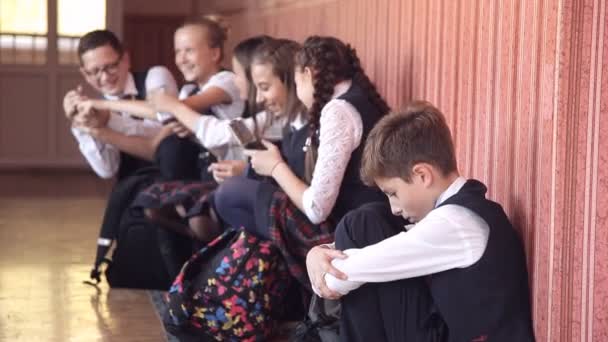Selectieve aandacht van ellendig schooljongen in uniforme zit alleen op de vloer in de hal van de school met hoofd op knieën huilen en eenzaam, voelt chatten en lachen klasgenoten in achtergrond — Stockvideo