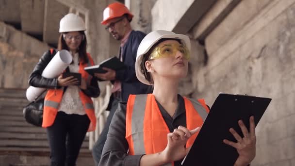 Uma mulher construtora ou arquiteta trabalha em um tablet e depois olha para a câmera. Ao fundo, conversando com os construtores — Vídeo de Stock