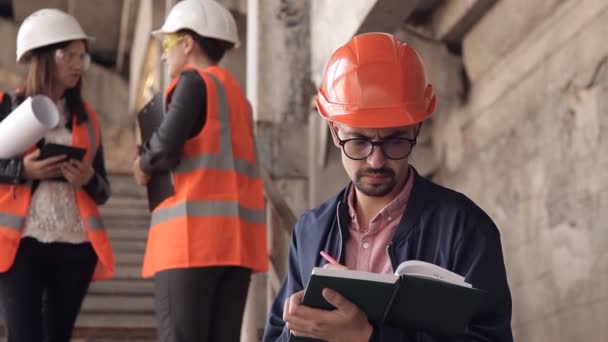 Muž stavitelé nebo architekt píše něco v poznámkovém bloku a pak se dívá na kameru. V pozadí, mluví s stavitelé — Stock video