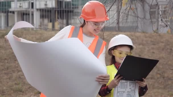 Zwei Kindermädchen in Bauhelmen, die ein weißes Blatt Papier betrachten oder zeichnen und lächeln — Stockvideo