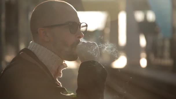 Homem de negócios de capa de chuva e óculos de sol fuma um cigarro na plataforma da estação ferroviária à espera de um trem — Vídeo de Stock