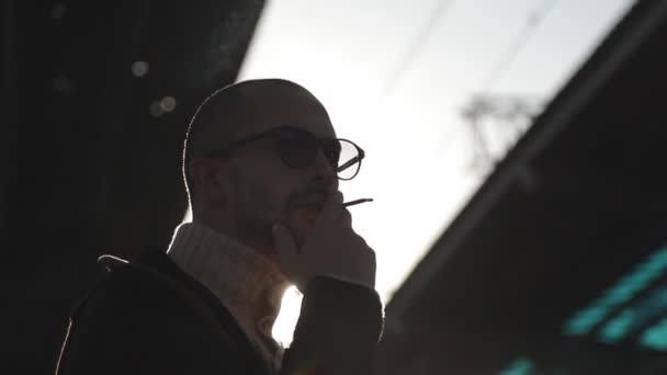 Hombre de negocios con impermeable y gafas de sol fuma un cigarrillo en la plataforma de la estación de tren esperando un tren — Vídeos de Stock