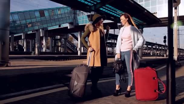 Two girls at the railway station with their suitcases out on the platform waiting for the train — Stock Video