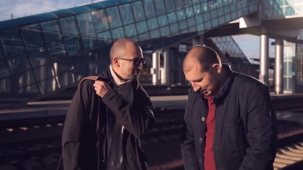 Двое мужчин на платформе железнодорожного вокзала ждут прибытия поезда и фотографируют селфи на смартфоне — стоковое видео