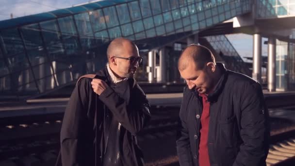 Two men on the platform of the railway station waiting for the arrival of the train and take pictures selfie on a smartphone — Stock Video