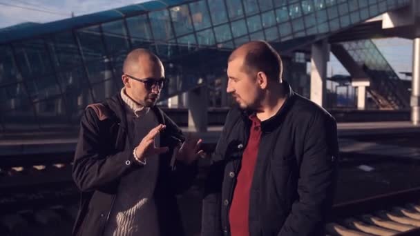 Dois homens na plataforma da estação ferroviária esperando a chegada do trem, conversando e animado sobre a viagem para a capital — Vídeo de Stock