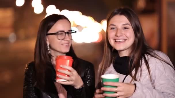 Två unga flickor gå genom gatorna i en obekant stad på en höst kväll och dricka kaffe — Stockvideo