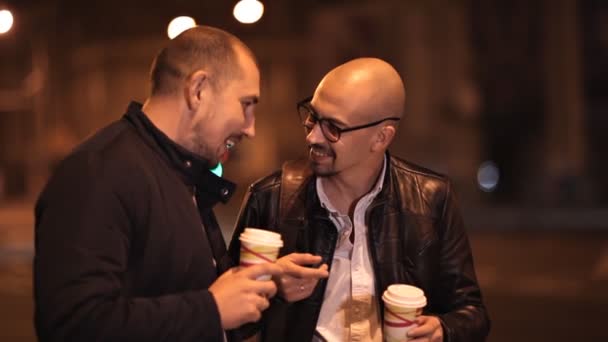 Two bald friends are walking in the evening in the city chatting, drinking coffee — Stock Video