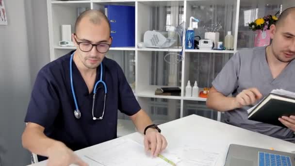 Jovens médicos sentam-se à mesa do escritório discutindo o tratamento e estudando as notas no caderno — Vídeo de Stock