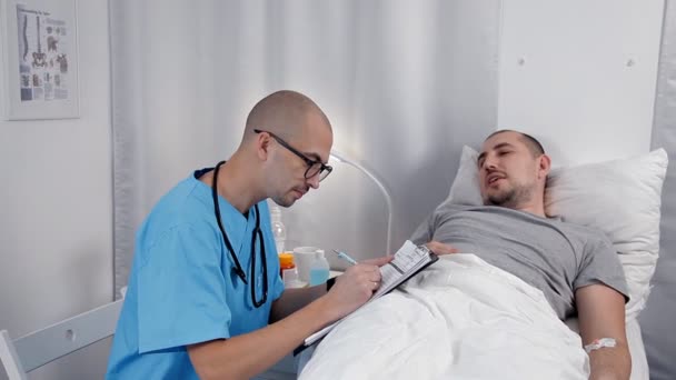 Paciente masculino joven hablando con un consultor en la sala de emergencias. El médico examina a un paciente y toma notas en un cuaderno — Vídeos de Stock