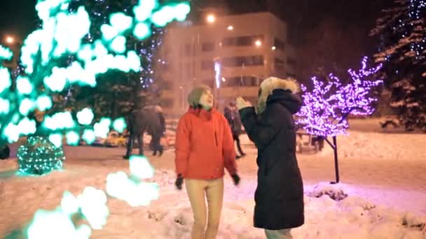 Дві молоді дівчата гуляють взимку через прикрашені вулиці міста. Новий рік, свято, сніг — стокове відео
