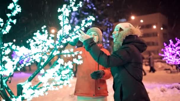 Dos jóvenes caminan en invierno por las calles decoradas de la ciudad. Año nuevo, vacaciones, nieve — Vídeos de Stock