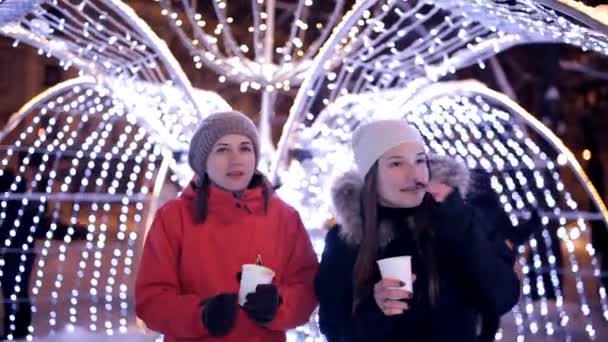 Молоді дівчата п'ють каву взимку в центрі міста, біля зимових прикрас. Різдво, Новий рік, свято — стокове відео