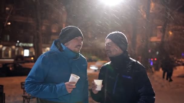 Двоє молодих чоловіків стоять на вулиці взимку, сильний сніг, розмовляють і п'ють каву — стокове відео
