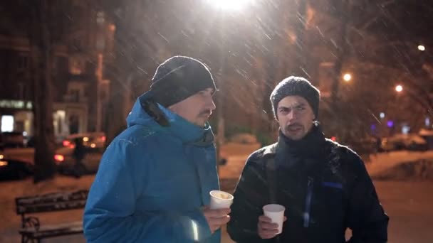 Zwei junge Männer stehen im Winter bei starkem Schneefall auf der Straße, unterhalten sich und trinken Kaffee — Stockvideo