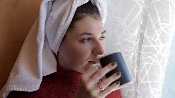 Vacker ung kvinna dricker kaffe och tittar genom fönstret medan du sitter på fönsterbrädan hemma — Stockvideo