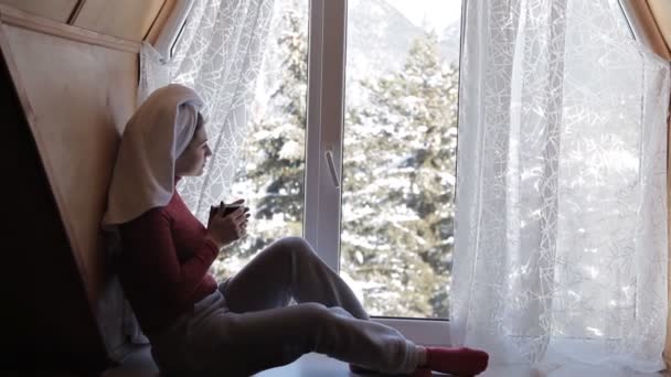 Bella giovane donna che beve caffè e guarda attraverso la finestra mentre seduto al davanzale della finestra a casa — Video Stock