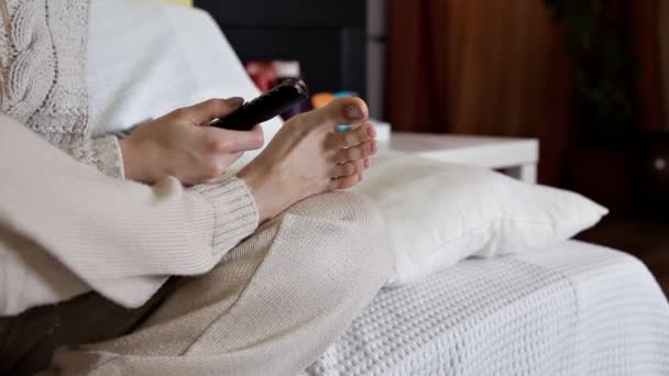 Girl sits barefoot at home and watches TV, switches channels — Stock Video