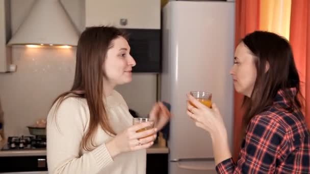Två unga flickor chattar i köket och dricker apelsinjuice medan middagen förbereds — Stockvideo