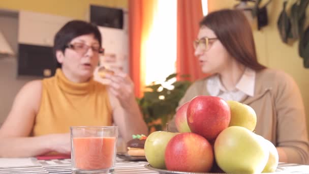 Mãe e filha com óculos bebem chá ou café na cozinha e comem bolos, falam, riem. Na placa de primeiro plano com maçãs — Vídeo de Stock