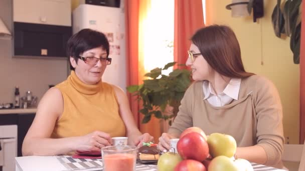 Mamá e hija pequeña con vasos beben té o café en la cocina y comen pasteles, hablan, ríen. En primer plano con manzanas — Vídeo de stock