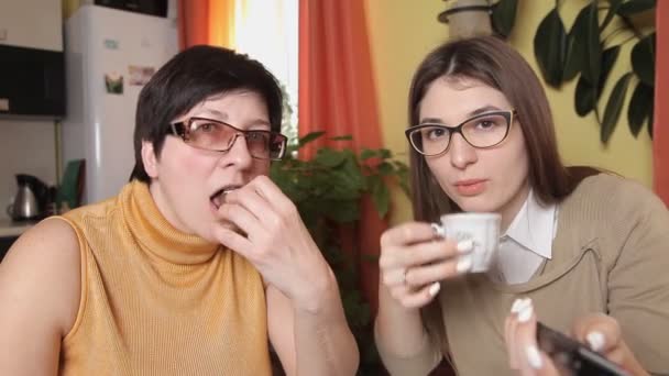 Mutter und Tochter mit Brille fernsehen, Tee trinken, Kaffee essen, Kuchen essen schalten die Fernbedienung und reagieren heftig. Blick in die Kamera — Stockvideo