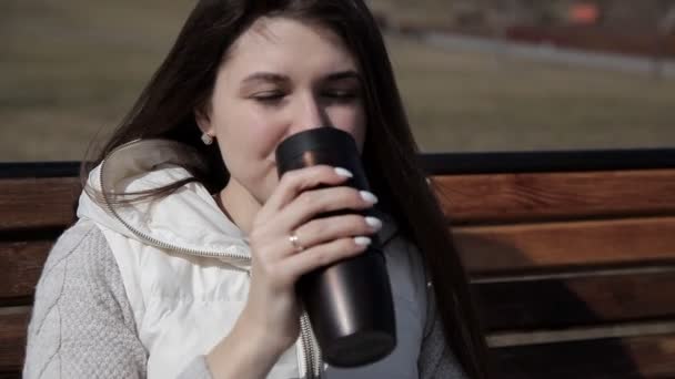 Chica en el parque en un banco bebiendo té, café de una taza termal. De cerca. gimiendo del sol — Vídeos de Stock