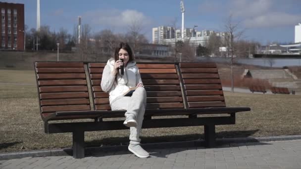 Una bella ragazza in tuta da ginnastica nel parco sulla panchina a leggere un libro e bere caffè da una tazza termica — Video Stock