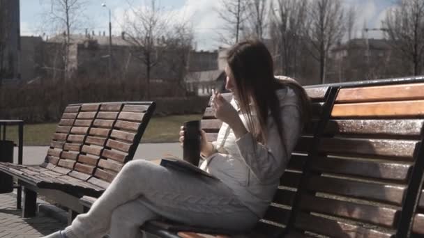 一个穿着运动服的美丽女孩在公园的长椅上, 坐在长凳上看书, 喝着热杯里的咖啡。喜剧的笑声 — 图库视频影像