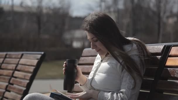 Uma menina bonita em um traje de treino no parque no banco lendo um livro e bebendo café de uma caneca térmica. Aponta um dedo e reconhece o lugar do livro — Vídeo de Stock