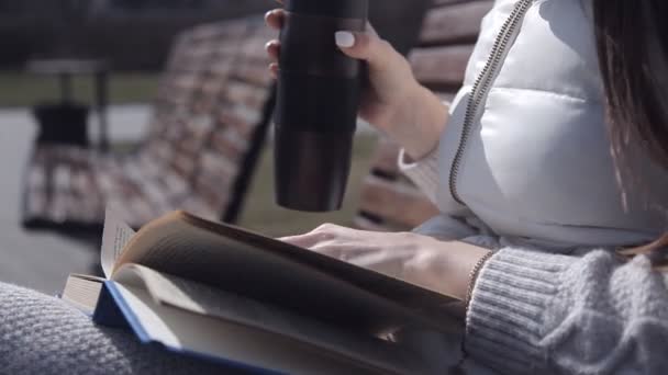 Una joven en el parque en un banco leyendo un libro y bebiendo café de una taza. de cerca — Vídeos de Stock